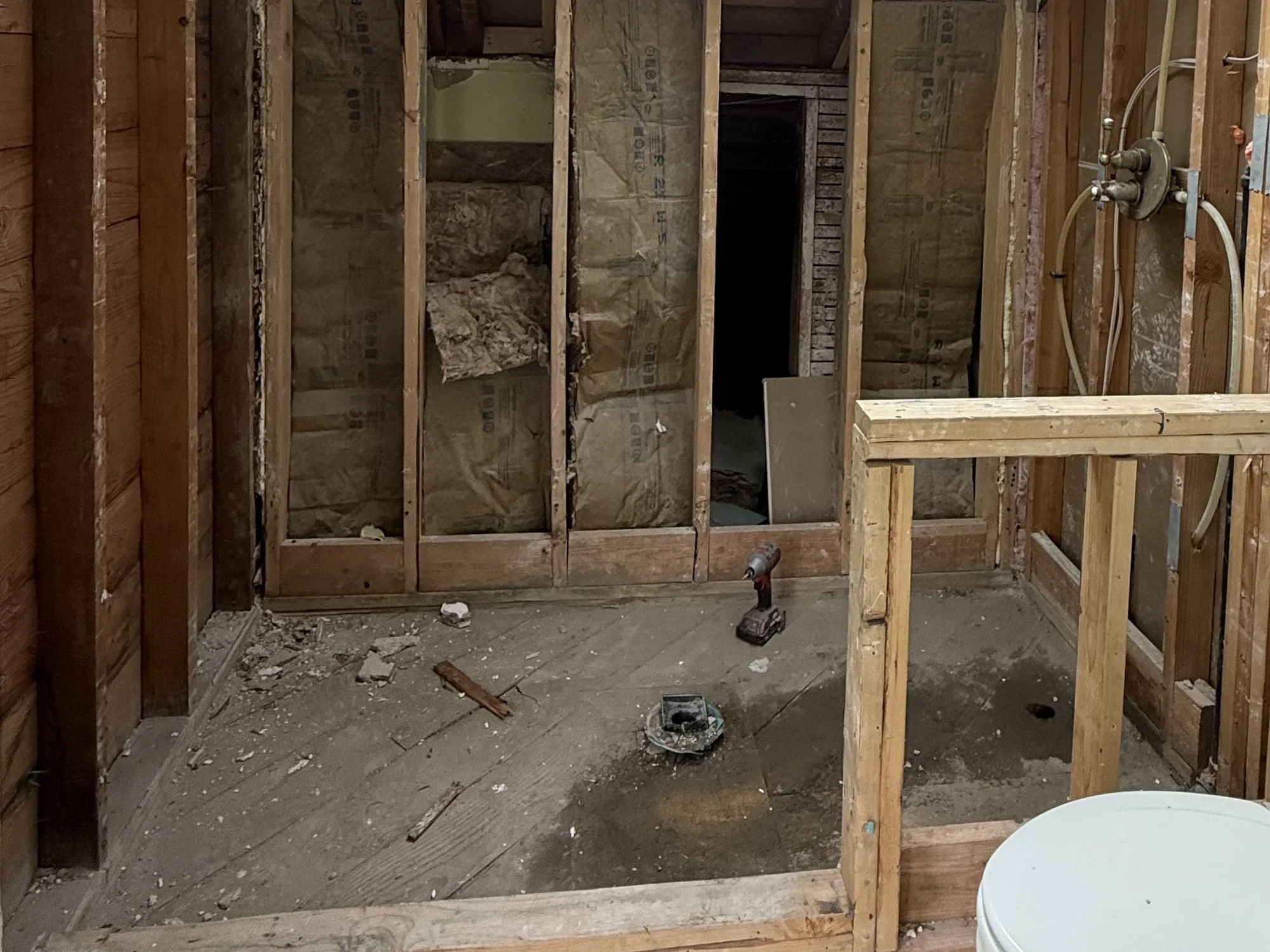 A bathroom stripped to bare studs and plank subfloor, with an old cast drain fitting standing open and a new stud wall framed on the right