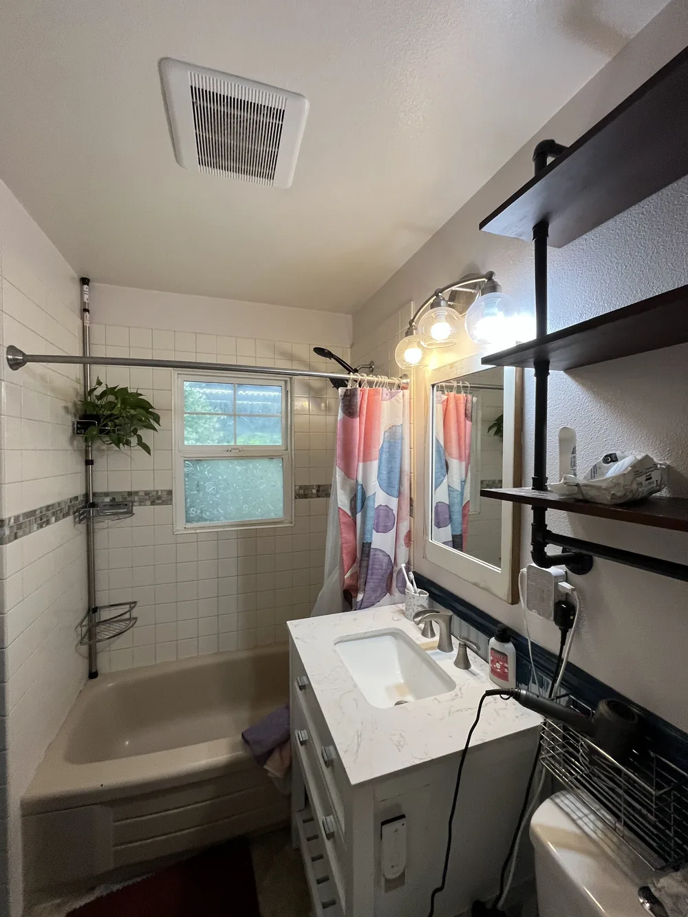 Bathroom before the remodel with a tub and shower curtain, a small vanity and open shelving on the wall