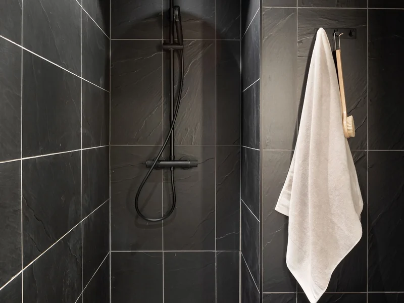 Dark slate look tile covering a shower corner, with a matte black bar valve, riser, handheld and hose, and a white towel on a black hook