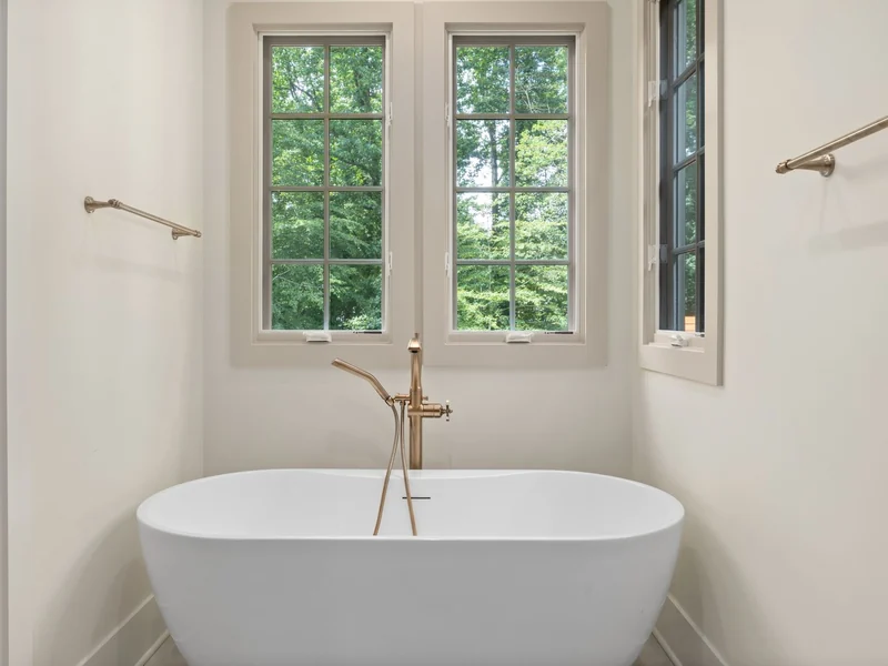 Freestanding oval tub under two windows with a floor-mounted brushed brass filler