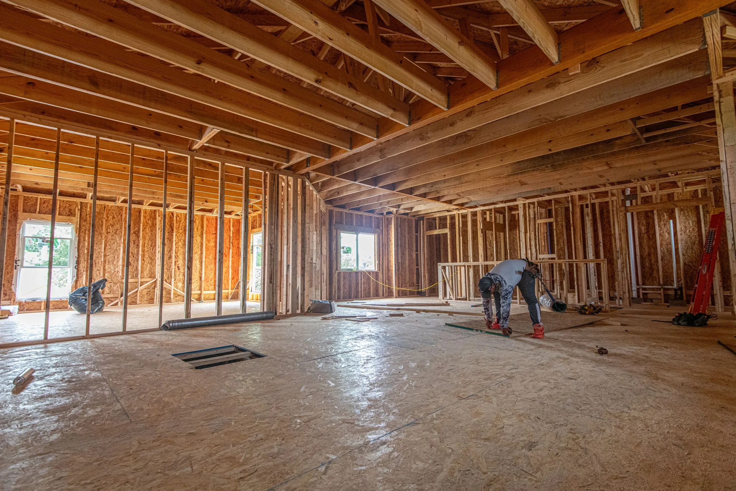 House at rough framing stage with open stud bays, exposed ceiling joists and a bare OSB subfloor, a worker bent over a level on the floor