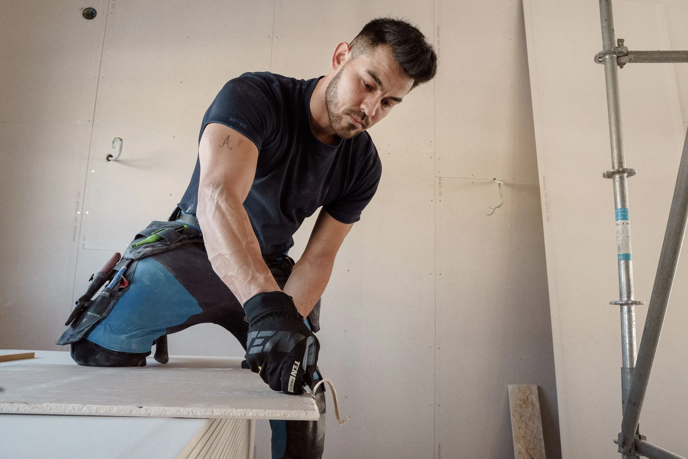 Worker in gloves and a tool belt lifting a drywall sheet from a stack, in a room with boarded untaped walls and a scaffold tower
