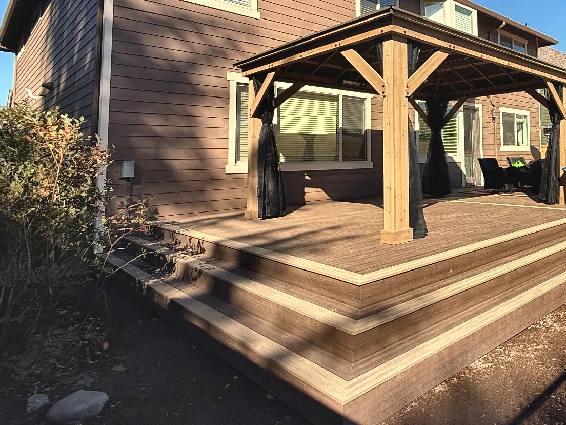 A raised deck with a lighter border board around its edge, two wide steps down to bare ground, and a four-post wood gazebo with dark mesh curtains set against a two-story house