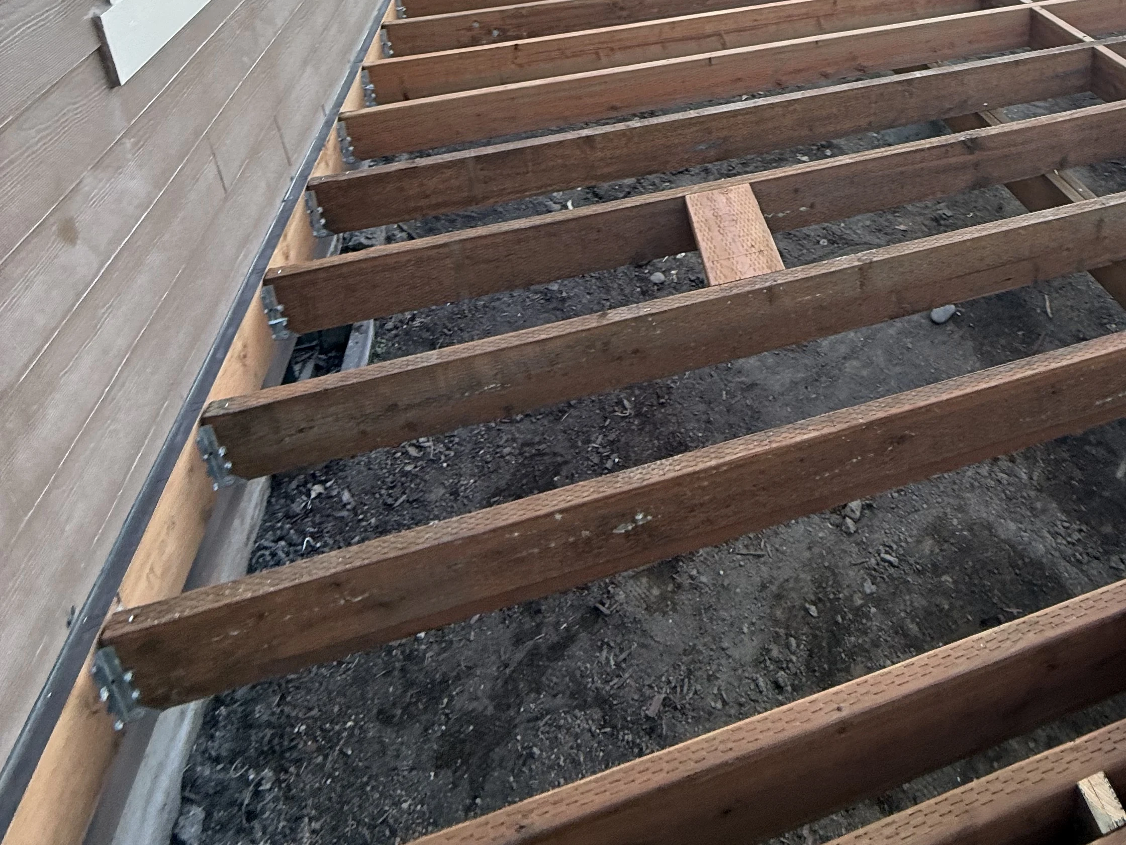 A ledger board fastened flat to the house siding, capped by a dark metal flashing strip, with galvanized joist hangers holding each deck joist