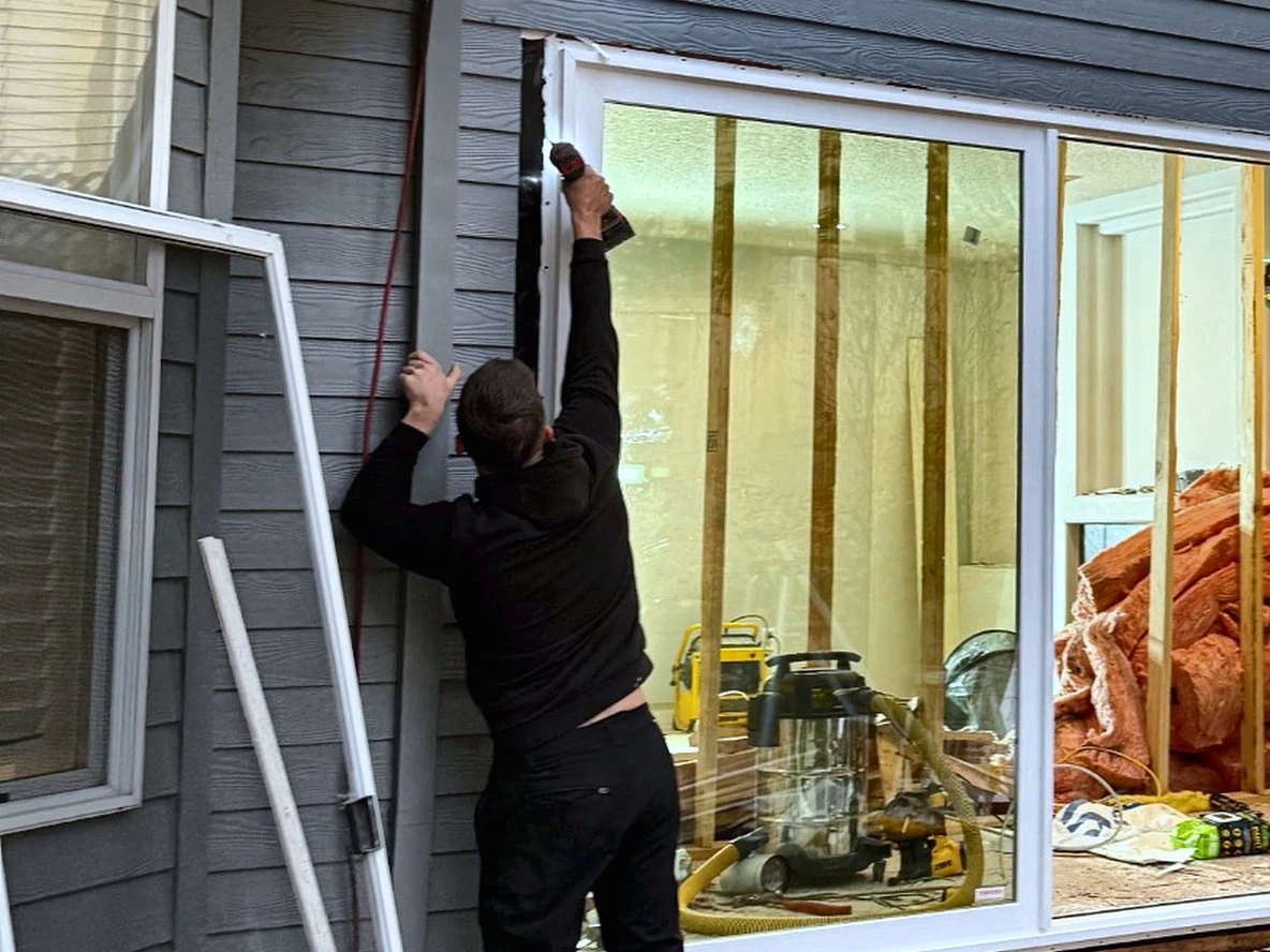 A large sliding door unit set into a cut opening in existing gray lap siding, with a black flashing membrane down the jamb and a worker driving a fastener through the white nailing fin
