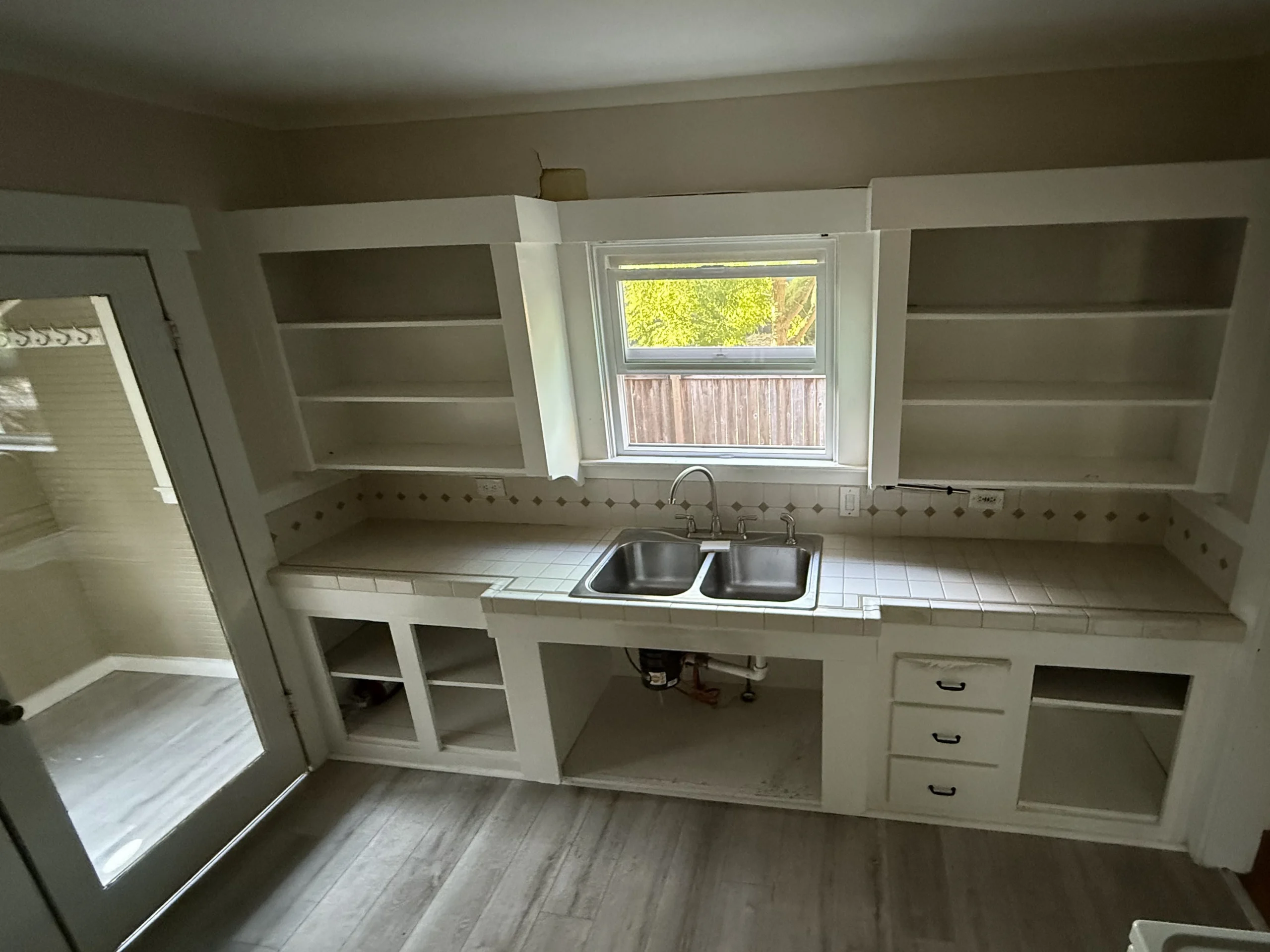 A kitchen with every cabinet door removed and bare shelves, a tile counter and diamond accent backsplash, a stainless double sink under a window, and the sink base open to the drain plumbing