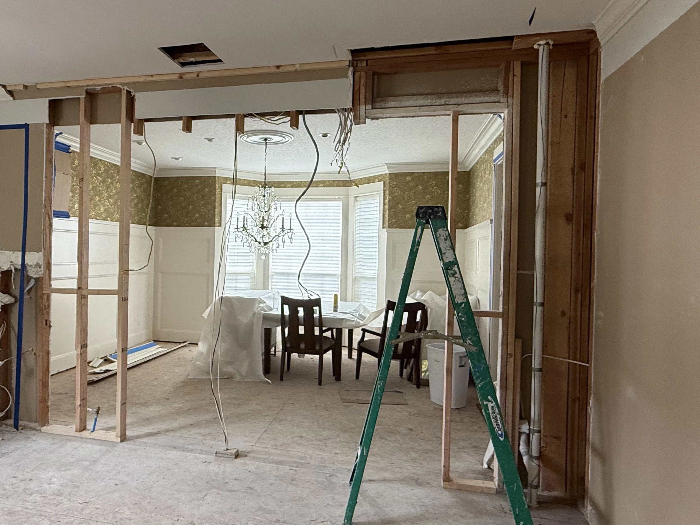 A horizontal wood member spans an opened interior wall above a vertical post on the right, with stripped studs and loose wiring hanging above, and beyond it a dining room with patterned wallpaper, white wainscot, crown molding, a chandelier and a table covered in plastic sheeting