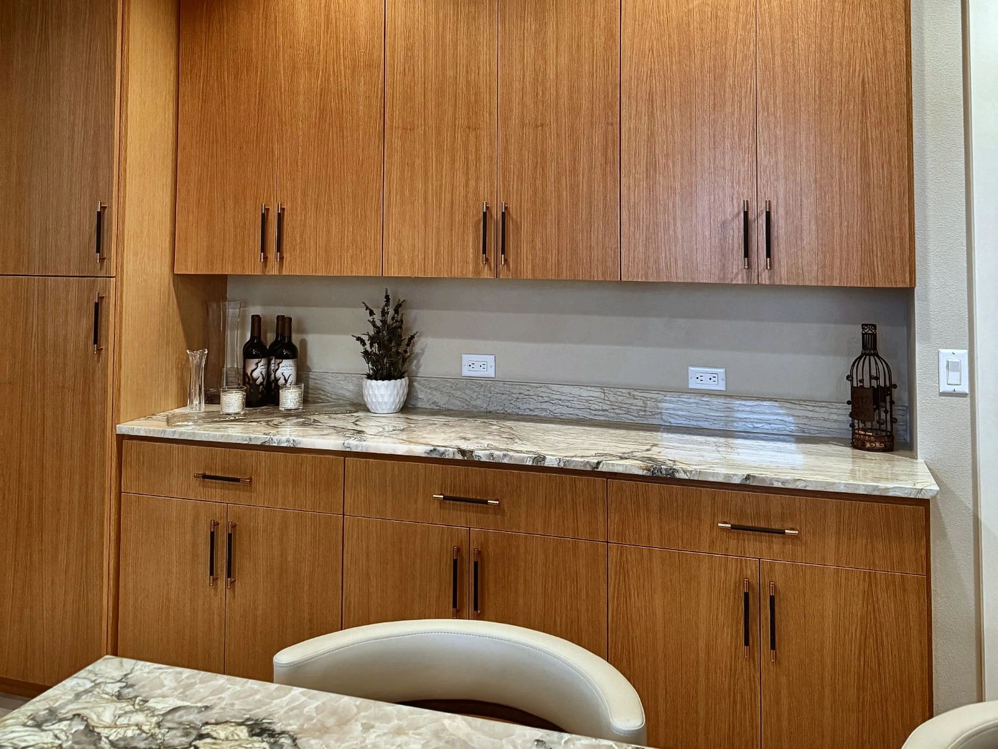 A wall of flat wood cabinets above and below a veined stone counter with a matching stone backsplash ledge and two outlets