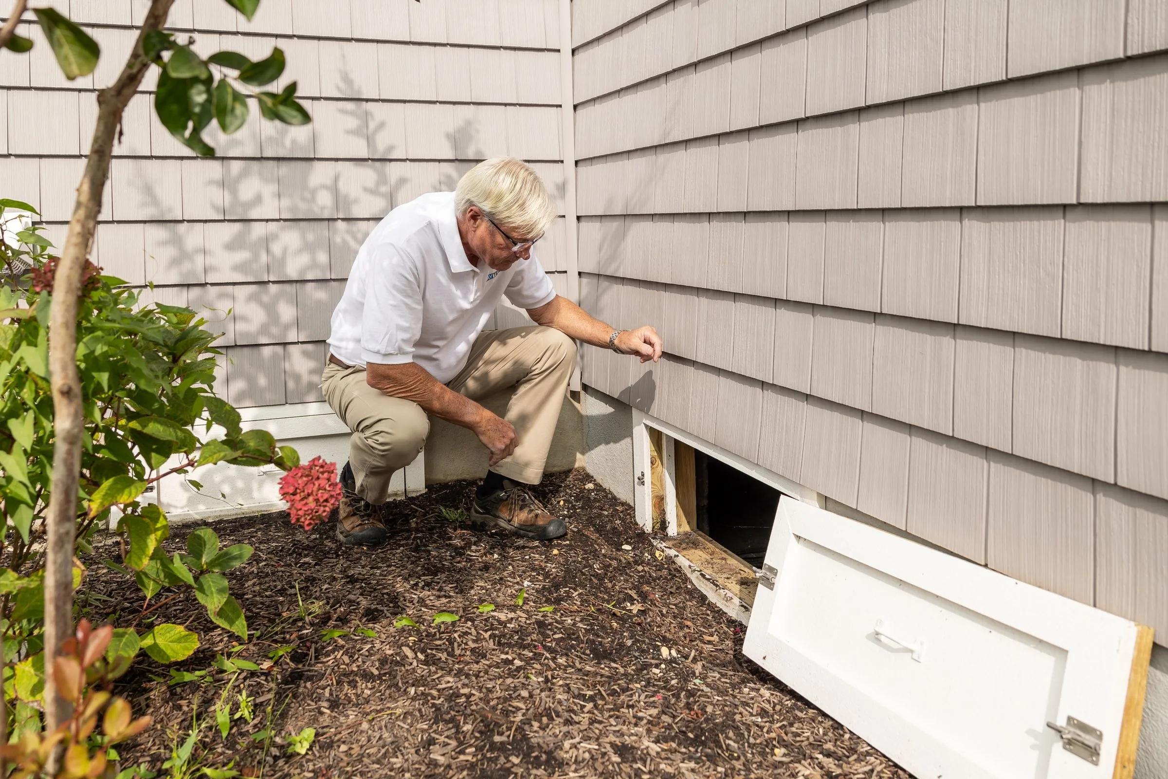 Man crouching in a mulch bed beside clean shake siding, looking into an open crawlspace vent with its white access door removed and leaning alongside