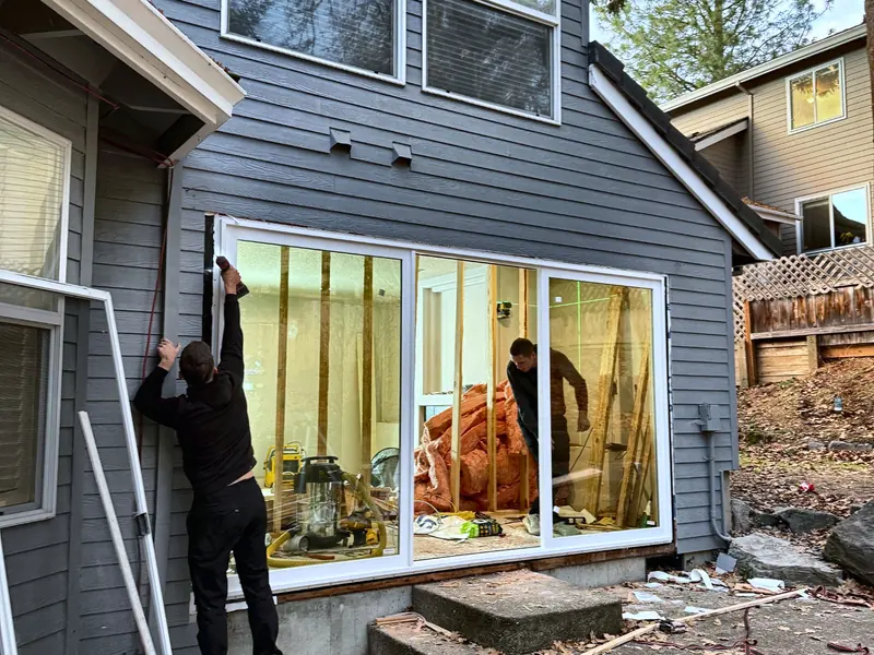 Two carpenters fitting a wide sliding glass door into the opening of a gray-sided house, with bare studs and insulation visible inside