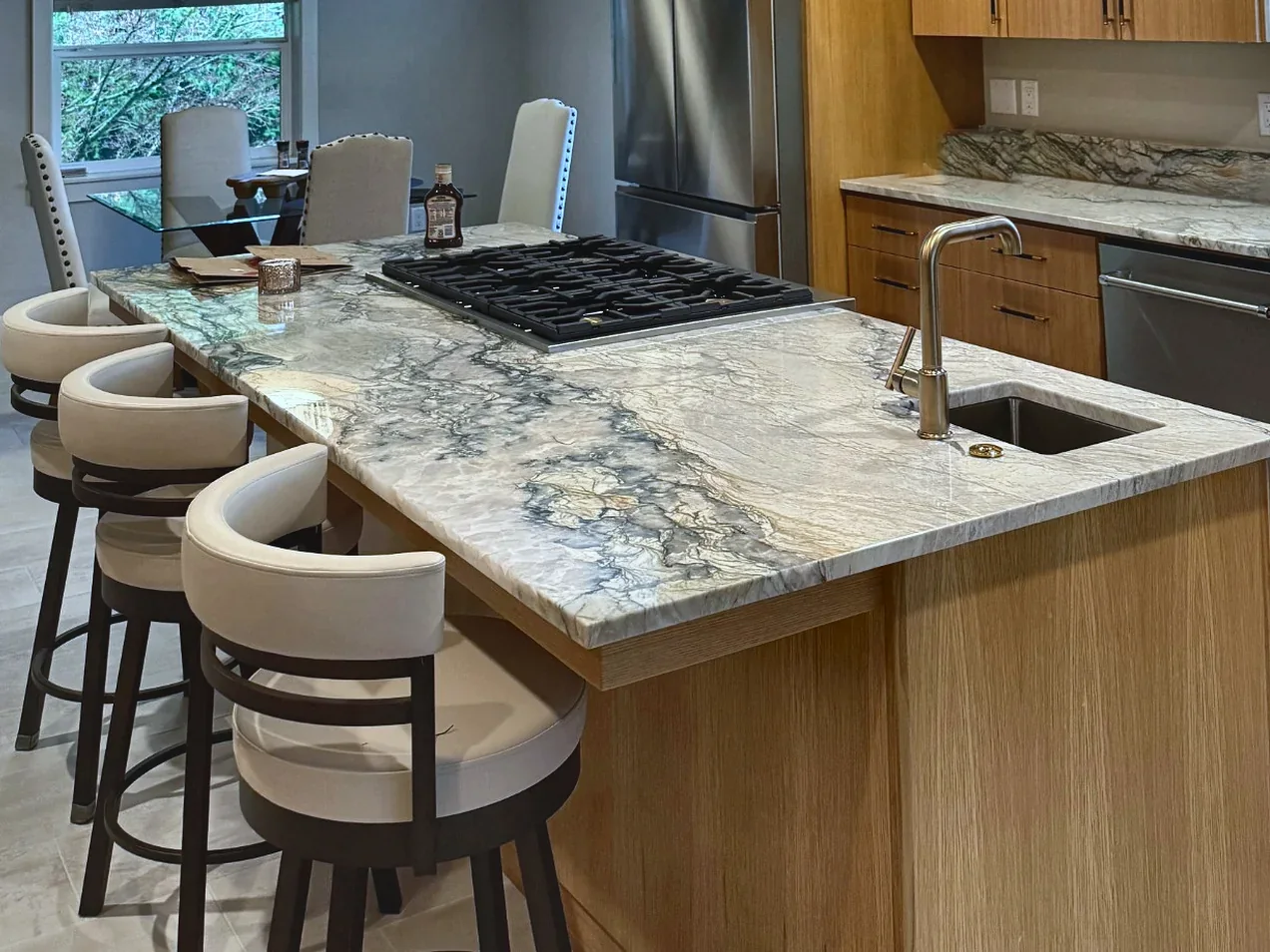 Remodeled kitchen with a veined stone island, four cream upholstered barstools, a gas cooktop, an undermount sink, warm wood flat-panel cabinets and stainless-steel appliances