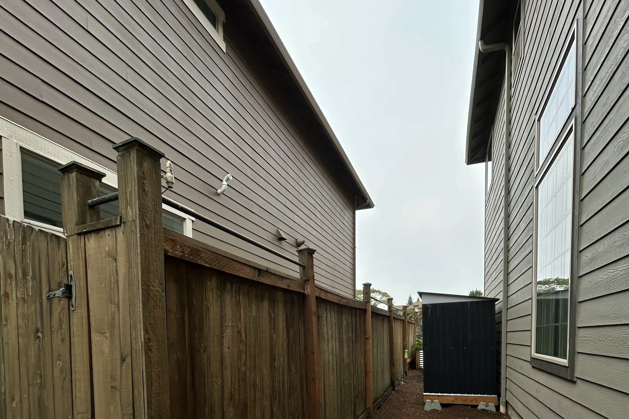 Narrow side yard between two two story homes with lap siding, shallow roof eaves, a weathered cedar fence and a metal storage shed under an overcast sky